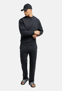 Black sweatshirt and joggers in soft fabric, featuring ribbed cuffs and subtle embroidery. Worn with a black cap and patterned slide sandals.
