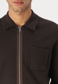 Jachetă maro cu fermoar, cu guler, având o buzunar frontal și un material texturat. Fermoarul de culoare argintie completează designul.