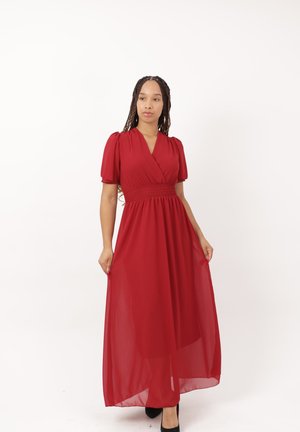 Donna con capelli intrecciati che indossa un lungo vestito rosso a maniche corte, in piedi contro uno sfondo bianco semplice.