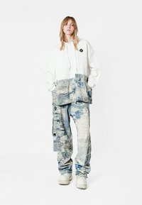 Sudadera blanca con una sección inferior de patrón azul y blanco, combinada con pantalones de corte suelto a juego. Presenta un logo de parche en el pecho.