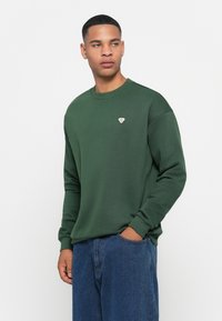Felpa verde con collo rotondo, polsini a costine e un piccolo logo ricamato sul petto, abbinata a jeans di denim.