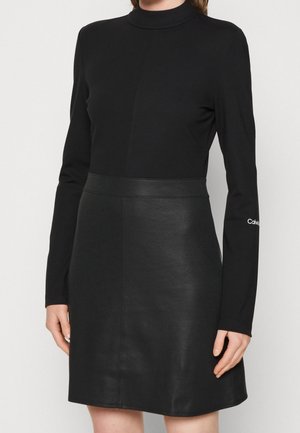 Musta mekko, jossa on korkea kaulus, pitkät hihat ja vartalonmyötäinen yläosa, joka siirtyy hieman flarettuun, nahkamaisiin hameeseen.