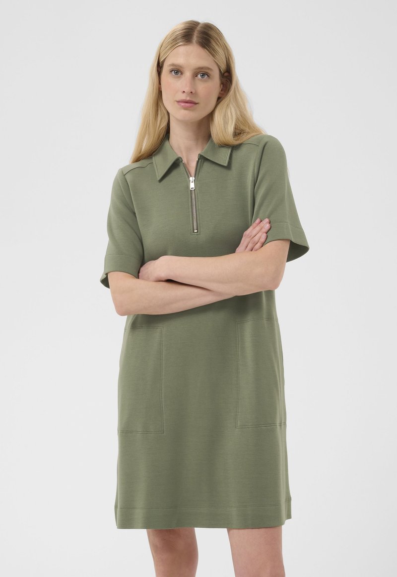 Femme aux longs cheveux blonds, portant une robe courte olive jusqu'aux genoux avec des manches courtes, des poches avant et un col zippé, bras croisés.