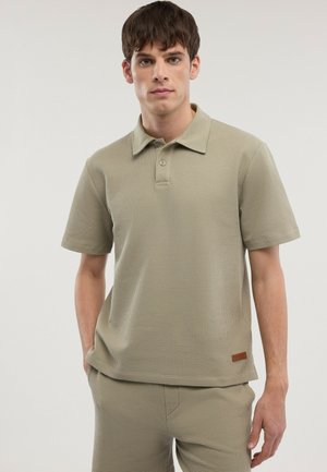 Mann trägt beige strukturiertes Polohemd mit kurzen Ärmeln und passende Hose, steht mit einer Hand in der Tasche vor einfachem Hintergrund.