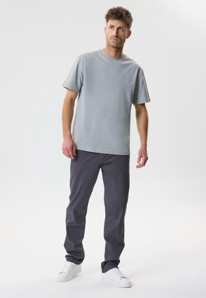 Noor mees, kellel on lühikesed pruunid juuksed, seljas helehall t-särk, tumehallid püksid ja valged tossud, seisab valgel taustal.