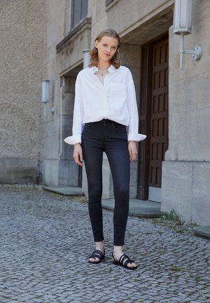 White oversized shirt with rolled cuffs, black skinny jeans, and black strappy sandals. Setting features textured stone walls and pavement.