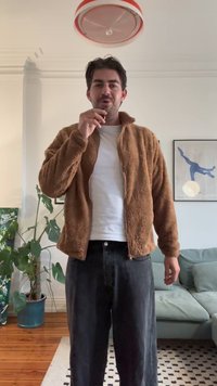 Braune Fleece-Jacke mit Reißverschluss und hohem Kragen, getragen über einem weißen T-Shirt, kombiniert mit lockeren schwarzen Jeans. Pflanze und Couch im Hintergrund.