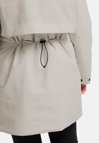 Parka impermeabile grigio chiaro con vita arricciata, dotata di cordoncino, chiusura a pressione e texture liscia e opaca.