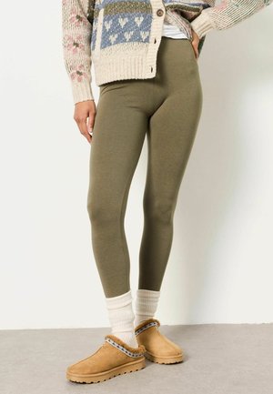 Persoon die olijfgroene legging draagt, een crèmekleurige gebreide trui met patronen, witte sokken en beige slip-on bontgevoerde schoenen, staande op een grijze vloer.