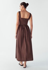 Rjava brezrokavna obleka s kvadratnim izrezom na hrbtu, prilegajočim vrhnjim delom in ohlapnim krilom. Material se zdi gladek z mehko teksturo.