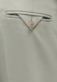 Κοντινό πλάνο ανοιχτό γκρι υφάσματος με μια μικρή τριγωνική φλόγα στερεωμένη με μπεζ κουμπί πάνω από μια οριζόντια σχισμή τσέπης.