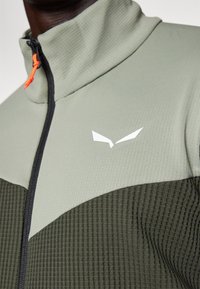 Veste légère zippée en vert et vert foncé, avec un tissu texturé, un col haut et un accent logo blanc contrastant.