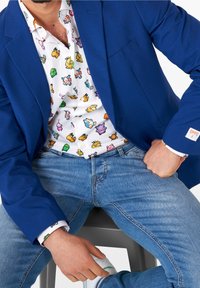Blazer blu sopra una camicia bianca con personaggi pixelati colorati, abbinato a jeans azzurri chiari e sneaker bianche.