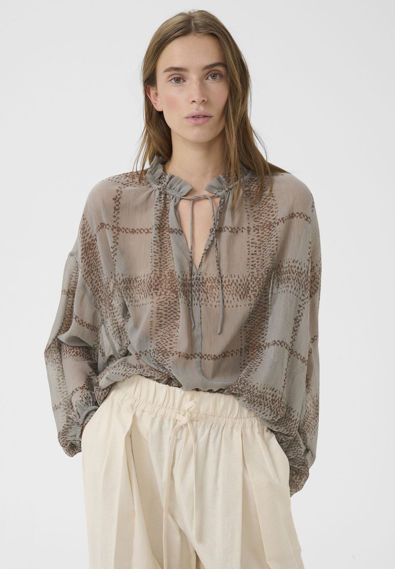 Femme portant un chemisier gris transparent et ample avec des motifs marron et un col à volants, associé à un pantalon crème taille haute à cordon de serrage.