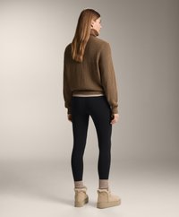 Rjav pleten pulover, črne legice in bež puhovke. Dolgi lasje, stoji od zadaj ob nevtralni ozadju.