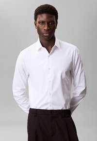 Hvid skjorte med lange ærmer og knaplukket krave, med en glat tekstur og en enkelt logodetalje på brystet, kombineret med sorte bukser.