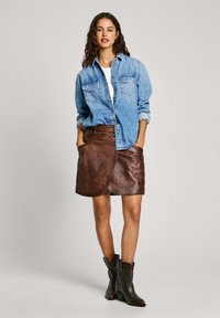 Denim srajca z gumbi, v kombinaciji s rjavim usnjenim zavitim krilom. Model nosi črne gležnjarje. Nevtralna ozadje.