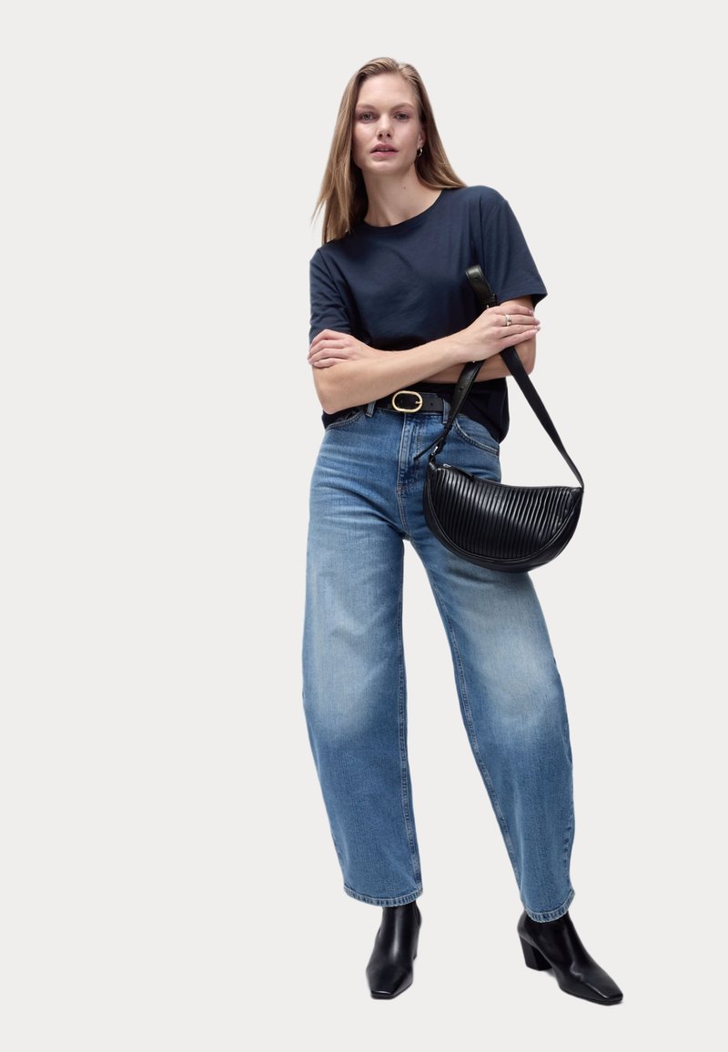 Svart ribbad läderaxelväska, marinblå t-shirt och ljusblå vida jeans. Modellen har på sig svarta klackstövlar med ankelcut och har armarna i kors.