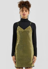 Frau in einem glitzernden goldenen Slipkleid über einem schwarzen Langarm-Turtleneck, mit hochgesteckten lockigen Haaren und hängenden Ohrringen.