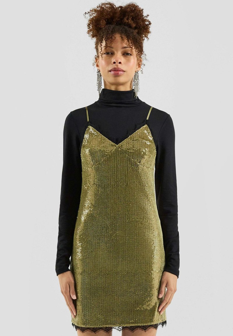 Frau in einem glitzernden goldenen Slipkleid über einem schwarzen Langarm-Turtleneck, mit hochgesteckten lockigen Haaren und hängenden Ohrringen.