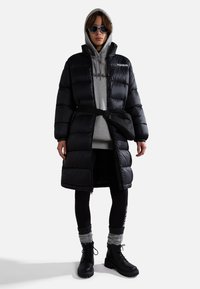 Persoon in een lange zwarte pufferjas, grijze hoodie, zwarte leggings, stevige zwarte laarzen, grijze beenwarmers, zwarte heuptas en donkere zonnebril.