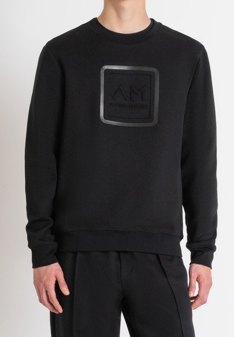 Personne portant un sweat-shirt noir à manches longues avec un logo carré en relief et un texte centré sur la poitrine.