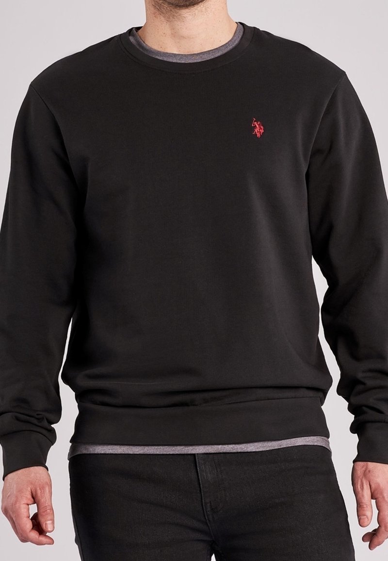 Homme portant un sweat-shirt noir à manches longues avec un logo rouge sur la poitrine, par-dessus un t-shirt gris et un pantalon noir, sur un fond uni.