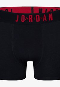 Schwarze Boxer-Briefs mit einem roten, elastischen Bund, auf dem "JORDAN" in Schwarz gedruckt ist. Glatte Textur mit sichtbaren Nähten auf der Vorderseite.