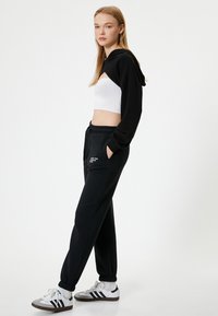 Schwarzer cropped Hoodie, weißes geripptes Tanktop, schwarze Jogginghose mit Taschen und weiße Sneaker mit schwarzen Streifen. Sauberer, lässiger Look.