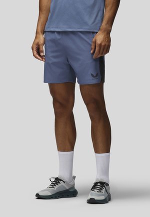 Homme portant un short de sport bleu, des chaussettes blanches et des chaussures de course grises, debout sur un fond gris.