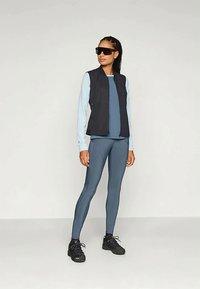 Black sleeveless vest over a light blue long-sleeve top, paired with dark blue leggings and black athletic shoes. Sunglasses complete the look.