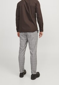 Muž stojící v hnědém dlouhém rukávu, šedých kostkovaných kalhotách a černých botách, odvrácený od jemného světlého pozadí.