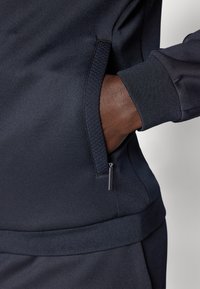 Marineblå genser med en jevn, matt tekstur. Har en sidelomme med glidelås og elastiske mansjetter for en tettsittende passform.