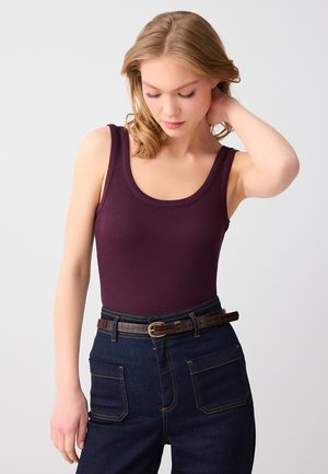 Vrouw met blond haar, gekleed in een mouwloos donkerpaars geribd topje en donkerblauwe spijkerbroek met een bruine riem, kijkt naar beneden en raakt haar nek aan.