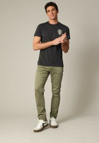 T-shirt en coton noir avec un col rond et un graphisme de lion, associé à un pantalon slim-fit vert olive et des baskets blanches avec des accents noirs.