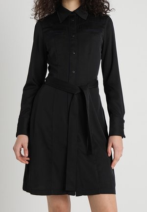 Mujer vistiendo un vestido camisa negro de manga larga con cuello, cierre de botones al frente, dos bolsillos en el pecho y un cinturón atado a la cintura.