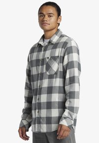 Camicia in flanella grigia e bianca con motivo a quadri, maniche lunghe, chiusura frontale con bottoni, taschino sul petto e orlo arrotondato.