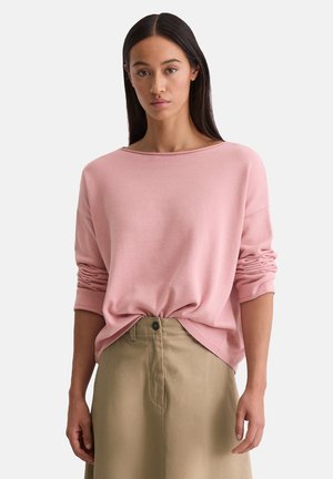 Pullover rosa chiaro con scollatura ampia, maniche lunghe e texture arricciata ai polsini. Abbinato a una gonna alta beige chiaro.