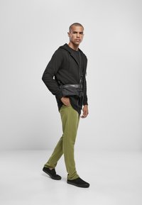 Sudadera negra con bolsillo frontal, pantalones de chándal verde oliva y zapatos negros sin cordones. El modelo lleva un bolso cruzado en la cintura.