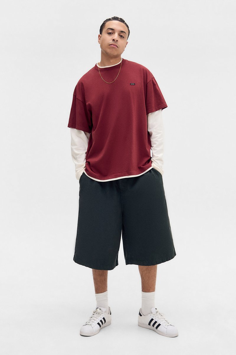 Rød t-skjorte med korte ermer lagdelt over en hvit langermet skjorte, svarte shorts og hvite sneakers med svarte striper. Enkelt design, avslappet passform.
