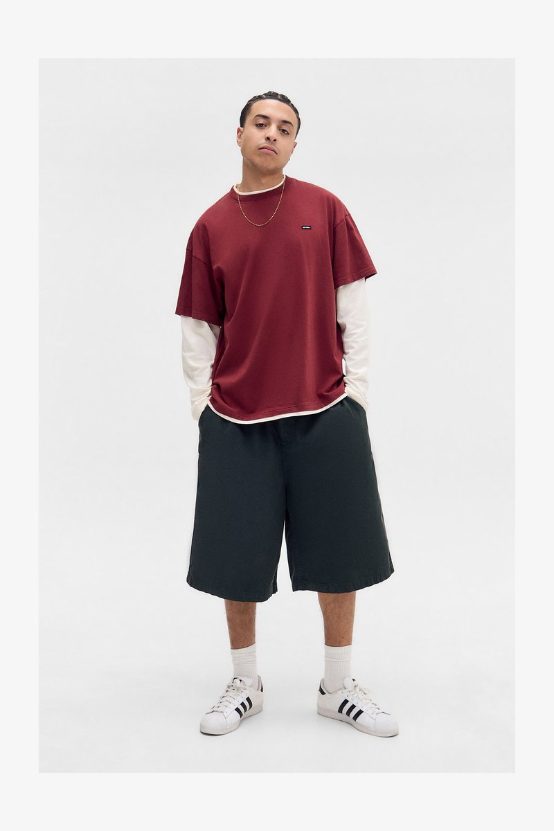Red short-sleeve t-shirt layered over a white long-sleeve shirt, black shorts, and white sneakers with black stripes. Simple design, relaxed fit.