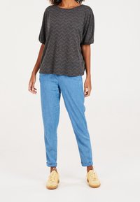 Žena v tmavě šedém volném topu se vzorem cik-cak, světle modrých ohrnutých džínách a žlutých teniskách, stojící na bílém pozadí.