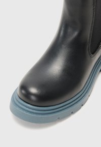 Bota de couro preta com bico arredondado e sola de borracha azul clara com detalhes texturizados. Inclui painéis laterais elásticos.