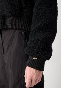 Pull noir en peluche avec des poignets côtelés et un accent de logo. La texture est douce, avec un design à poils épais. Associé à un pantalon sombre.