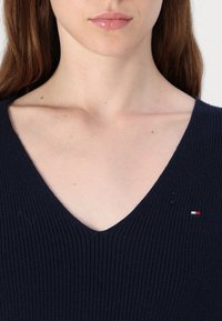 Gros plan sur le cou et la partie inférieure du visage d’une personne portant un pull col en V côtelé bleu marine avec un petit logo rouge, blanc et bleu.