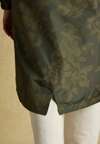 Tamsiai žalia gėlėmis papuošta striukė su šoniniu įpjovimu, vilkima virš baltų kelnių, matosi ranka su sidabriniu žiedu kairėje pusėje.