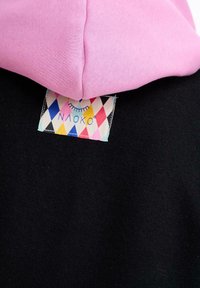 Ružová kapucha preložená cez čiernu látku s farebnou látkovou nálepkou s diamantovým vzorom, ktorá obsahuje dizajn zatvoreného oka a text.