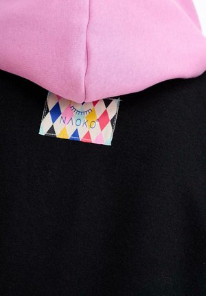 Różowy kaptur złożony na czarnym materiale, z kolorową metką odzieżową w diamentowy wzór, zawierającą zamknięte oko i tekst.