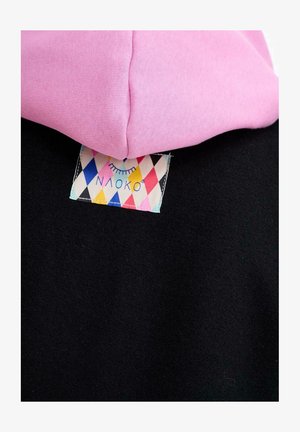 Różowy kaptur złożony na czarnym materiale, z kolorową metką odzieżową w diamentowy wzór, zawierającą zamknięte oko i tekst.