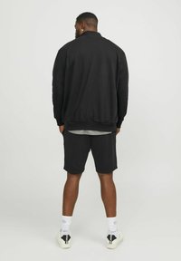 Sudadera negra con ribetes en el dobladillo y en los puños, combinada con pantalones cortos negros. Modelo con zapatillas blancas y calcetines deportivos a rayas. Fondo liso.